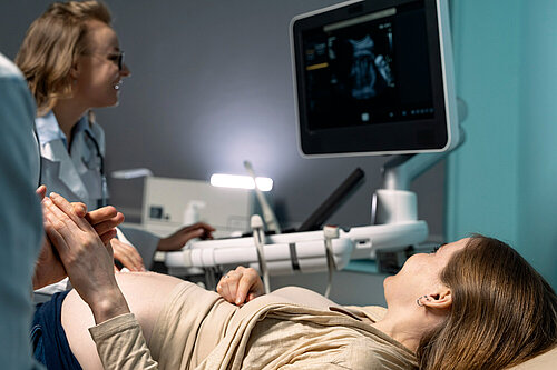 In the foreground woman is lying down in a medical setting, looking up at a screen showing an ultrasound. On the left of the photo, is a nurse operating the ultrasound.